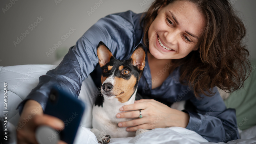 Beautiful young adult woman in pajamas doing selfie on smartphone with her dog bassengi in the morning in bed