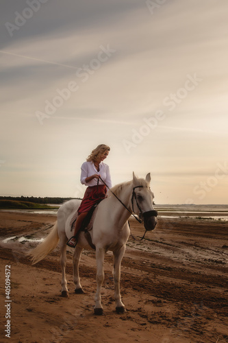 Cute happy young woman on horseback in summer beach by sea. Rider female drives her horse in nature on evening sunset light background. Concept of outdoor riding, sports and recreation. Copy space