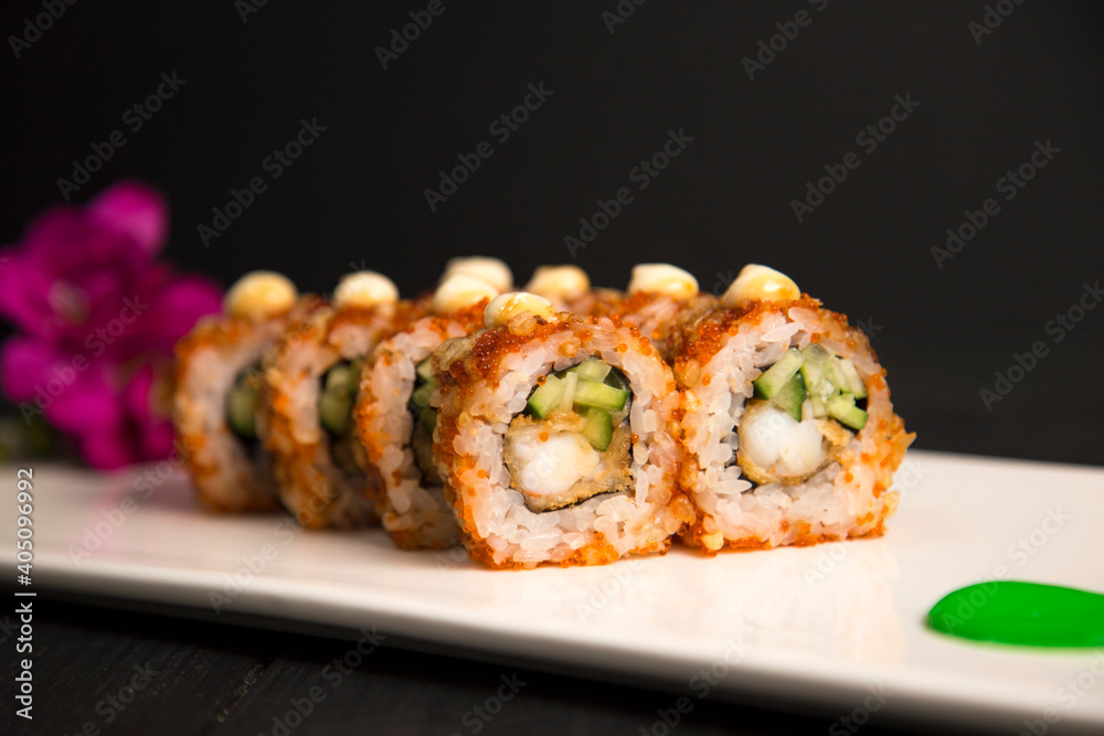 Macro shot of crunchy Japanese sushi roll with tempura black tiger shrimp (prawn), cucumber served with sauce on top. Purple flower, green painting brush stroke on white plate, Black Wooden background