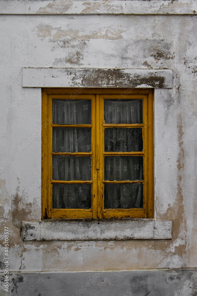 Detail of old traditional house in a Portuguese village with yellow wooden window and embroidered curtains. Modest house with dirty damaged white walls and rustic window frame