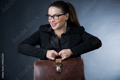 Donna d'affari bionda con occhiali da vista, vestita con  tailleur nero e felice con la sua borsa in pelle , isolata su sfondo grigio photo