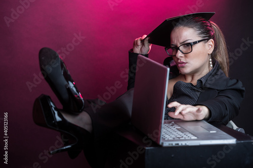 Manager donna con i capelli raccolti  con occhiali da vista, vestita con  tailleur nero lavora al computer con i piedi sopra il tavolo , isolata su sfondo rosso