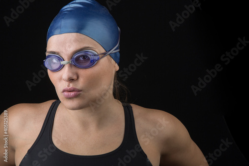 Giovane ragazza bianca con costume da bagno nero, cuffietta blu e occhialini da nuotatrice, guarda con aria decisa e concentrata prima della gara, isolata su sfondo nero . photo