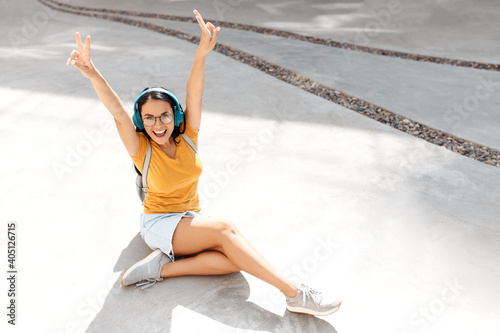 Excited brunette young woman student listening music on headphones sitting on cement floor in front of rusty wall.