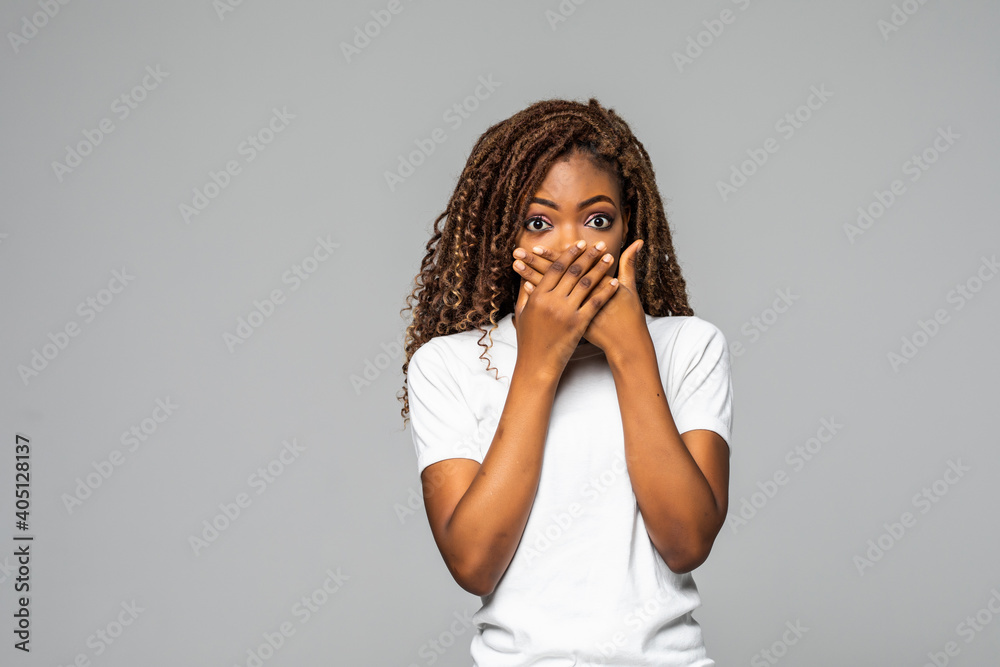 Shocked young African woman covering mouth with hands and looking at camera while standing against white background Shocked young African woman covering mouth with hands and looking at camera while standing against white background