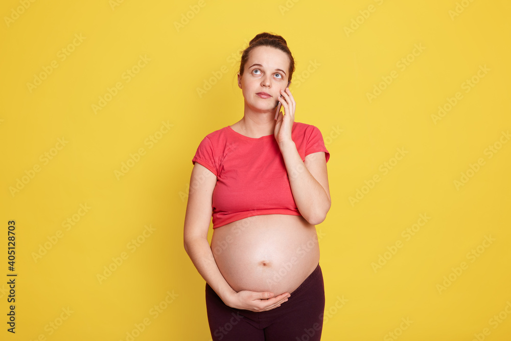 Smiling pregnant woman talking on her smart phone while posing isolated over yellow background, touching her bare tummy and looking away with dreamily expression. Smiling pregnant woman talking on her smart phone while posing isolated over yellow background, touching her bare tummy and looking away with dreamily expression.