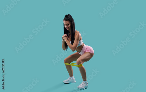 Portrait of a fitness woman doing squats with elastic band on blue background. Sporty girl squatting