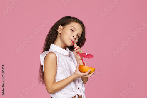 cute little child girl holding fresh juicy grapefruit and drinks juice isolated on pink background