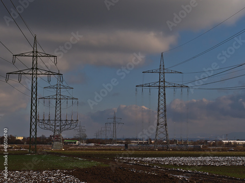 Power pylons (also transmission towers) and overhead power lines above agricultural fields covered by residual snow in winter season neat Stuttgart, Germany on cloudy day.