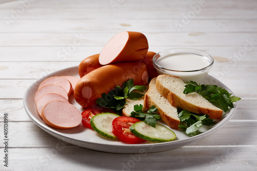 Sausages with vegetables and herbs garnish with sauces and bread on a gray ceramic plate on a white wooden table.