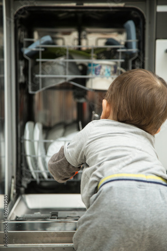  Little child boy toddler helping mom to disassemble the dishwasher. The dishwasher is open, the child is sitting next to him and holding clean dishes in his hands.