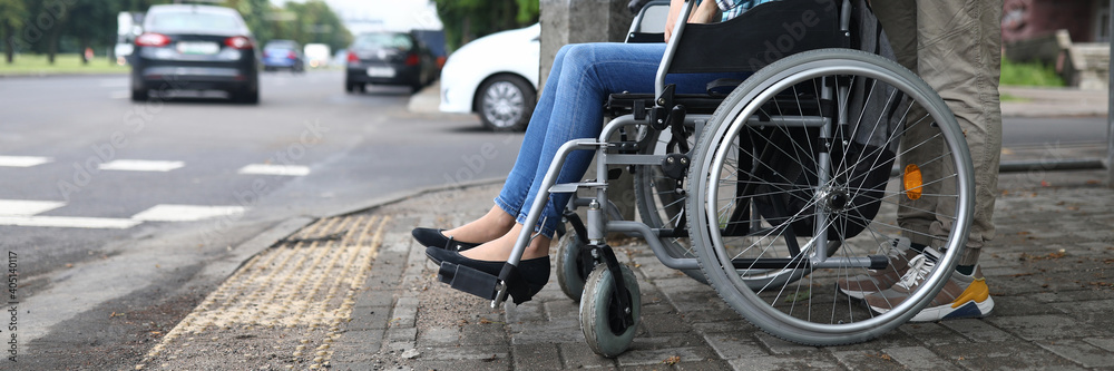 Close-up of man accompany female in wheelchair. Person taking care of lover. Carefully crossing street. Lady in jeans and blue shirt. Walk in city. Disabled people concept Close-up of man accompany female in wheelchair. Person taking care of lover. Carefully crossing street. Lady in jeans and blue shirt. Walk in city. Disabled people concept