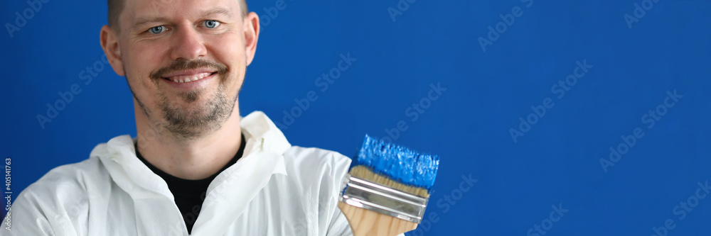 Portrait of paintbrush covered with blue paint. Smiling male worker in uniform on wall background. Professional interior designer. Renovation and construction site concept Portrait of paintbrush covered with blue paint. Smiling male worker in uniform on wall background. Professional interior designer. Renovation and construction site concept