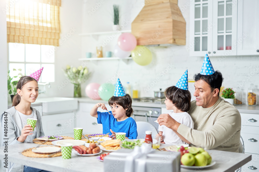 Little party. Loving middle aged latin father with children wearing birthday caps on heads and having a dinner while celebrating birthday together at home