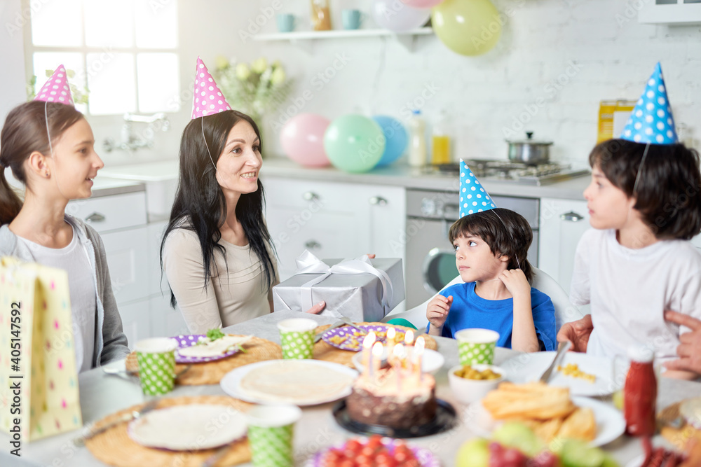 Family party. Cheerful middle aged latin mother holding gift box, talking to her lovely children while having dinner, celebrating birthday together at home