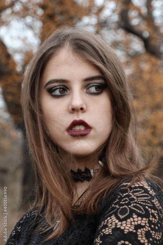 The face of a young witch. Makeup in the Gothic style. Spooky atmosphere in the autumn garden. Brunette with painted lips. The gaze is set aside. Black clothes. Shooting for Halloween. The face of a young witch. Makeup in the Gothic style. Spooky atmosphere in the autumn garden. Brunette with painted lips. The gaze is set aside. Black clothes. Shooting for Halloween.