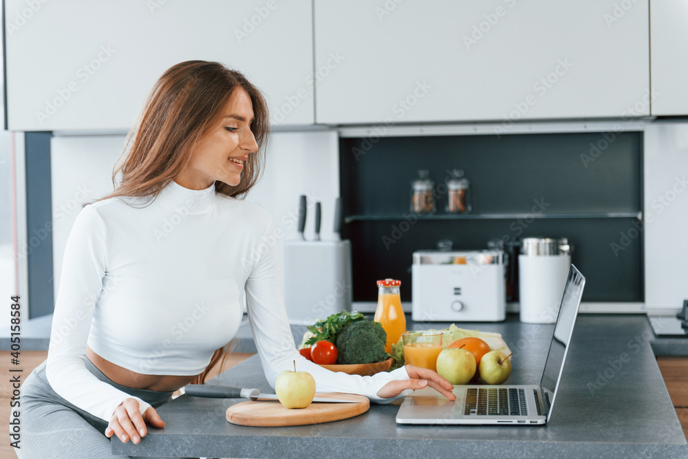 Uses laptop. Young european woman is indoors at kitchen indoors with healthy food