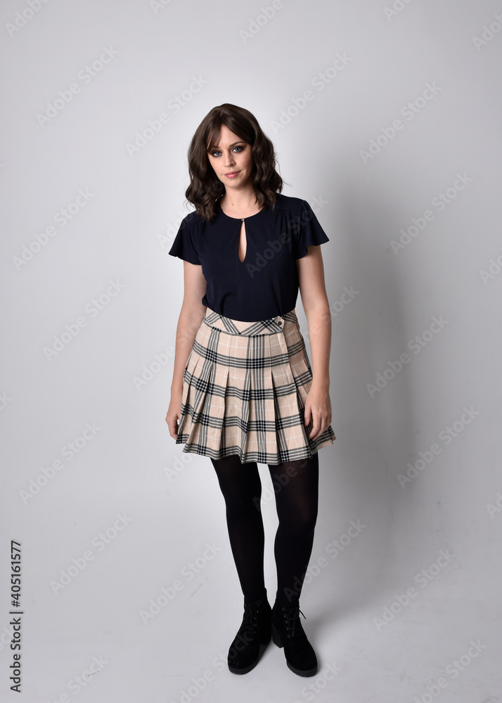 Full length portrait of pretty brunette woman wearing tartan skirt and blouse. Standing pose on the ground, against a studio background. Full length portrait of pretty brunette woman wearing tartan skirt and blouse. Standing pose on the ground, against a studio background.