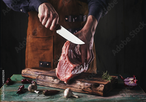A guy in a leather apron is slicing raw meat. The butcher cuts the pork ribs. Meat with bone on a wooden cutting board..
