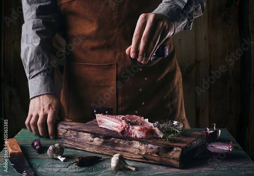 A guy in a leather apron sprinkles black pepper on a steak with a bone. Raw meat on a wooden cutting board..