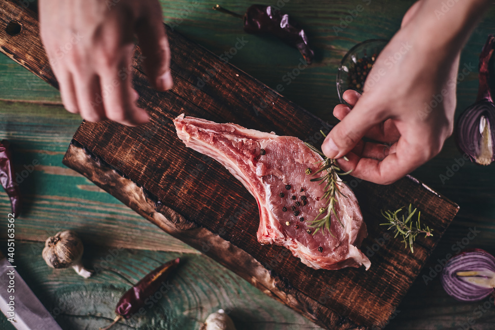 Pork steak with bone on a wooden cutting board. Man's hands lay a rosemary branch on a piece of meat..