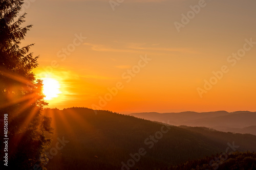 Sun setting over a horizon of orange hills with a coniferous tree on the left and clouds moving in the background over the Beskydy countryside. © Lukas