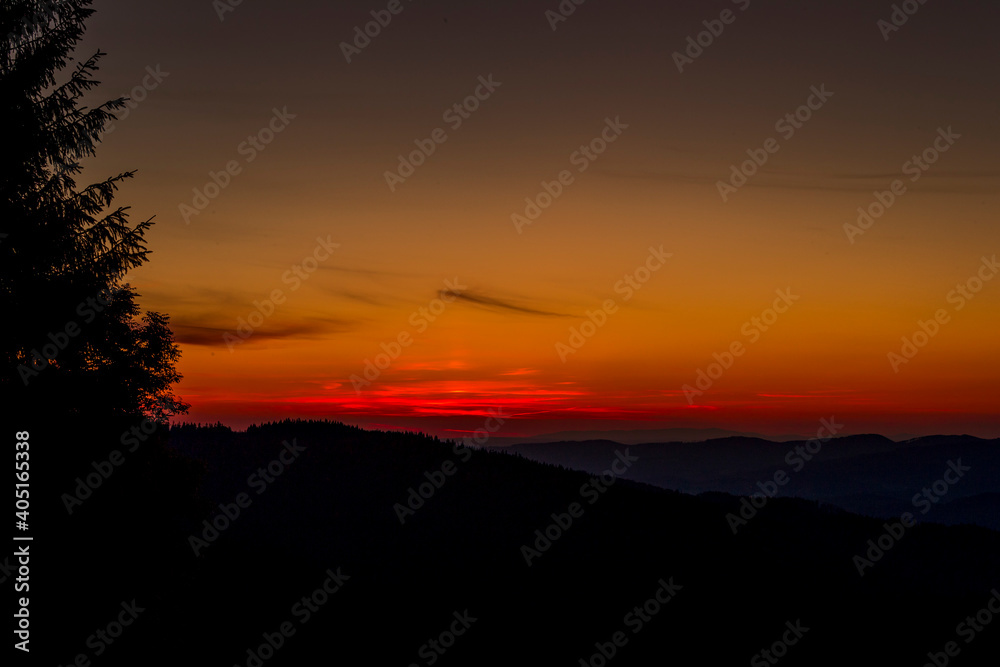 Sun setting over a horizon of orange hills with a coniferous tree on the left and clouds moving in the background over the Beskydy countryside.