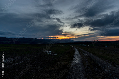 A dirt road leading around a field during an orange sunset on the horizon of the setting sun behind the hills.