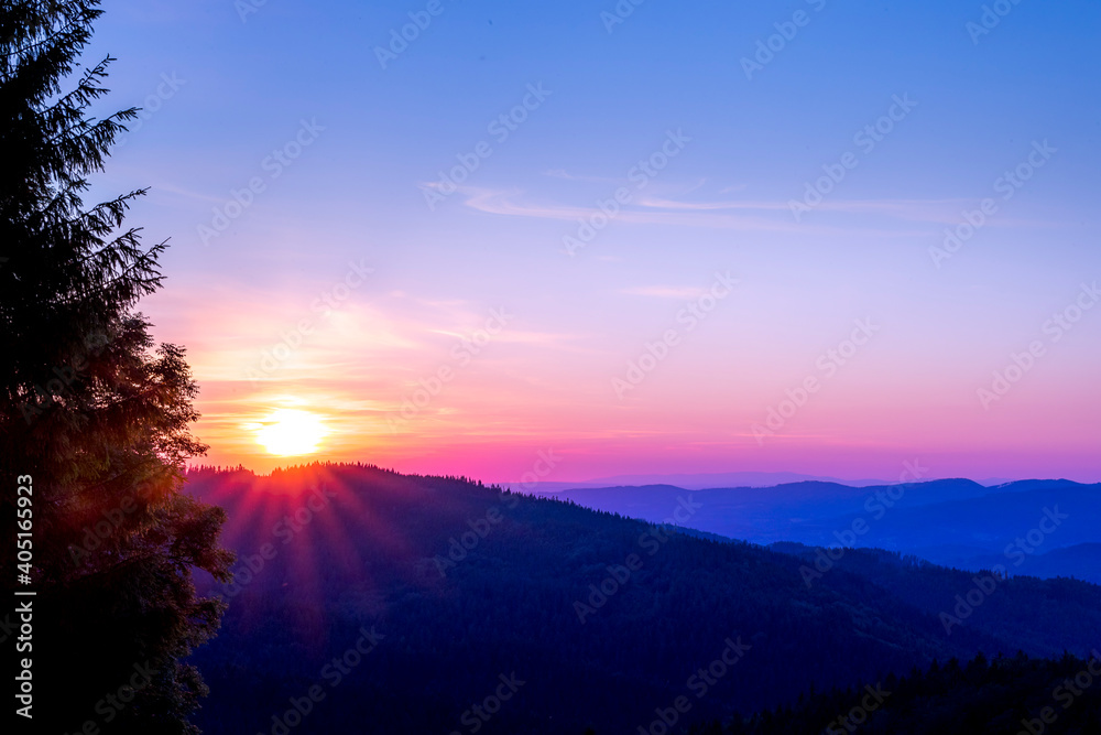 Sun setting over a horizon of orange hills with a coniferous tree on the left and clouds moving in the background over the Beskydy countryside.