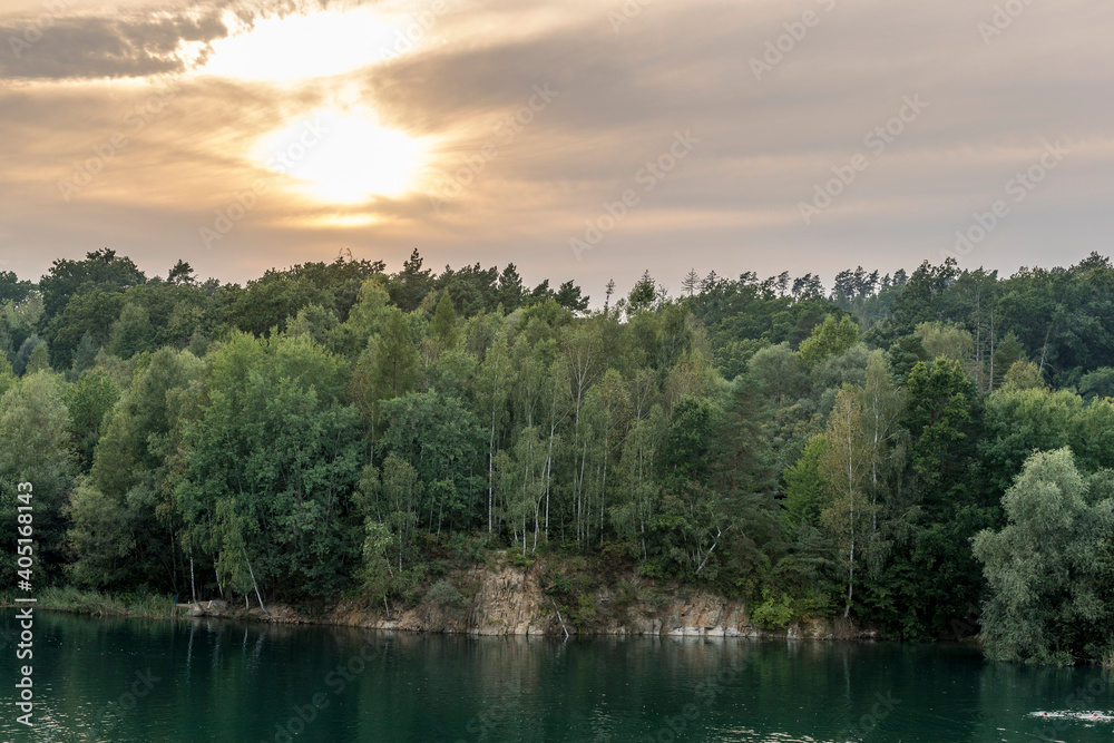 Flooded quarry and trees lying on the shore during sunset with fast moving clouds and a blue-orange sky in the background.