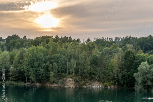 Flooded quarry and trees lying on the shore during sunset with fast moving clouds and a blue-orange sky in the background.