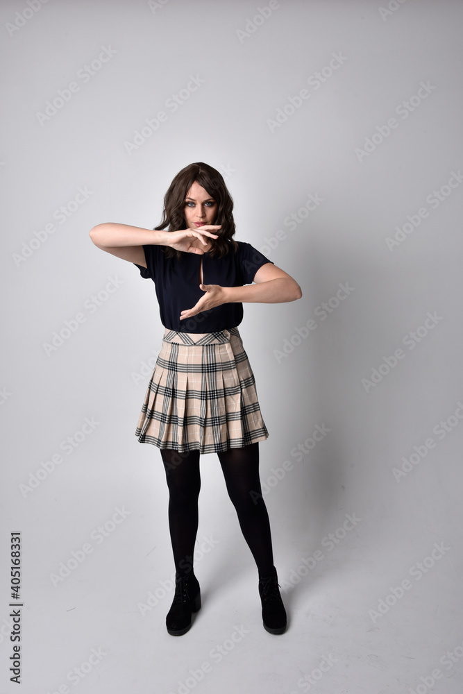Full length portrait of pretty brunette woman wearing tartan skirt and blouse.  Standing pose on the ground,  against a  studio background.