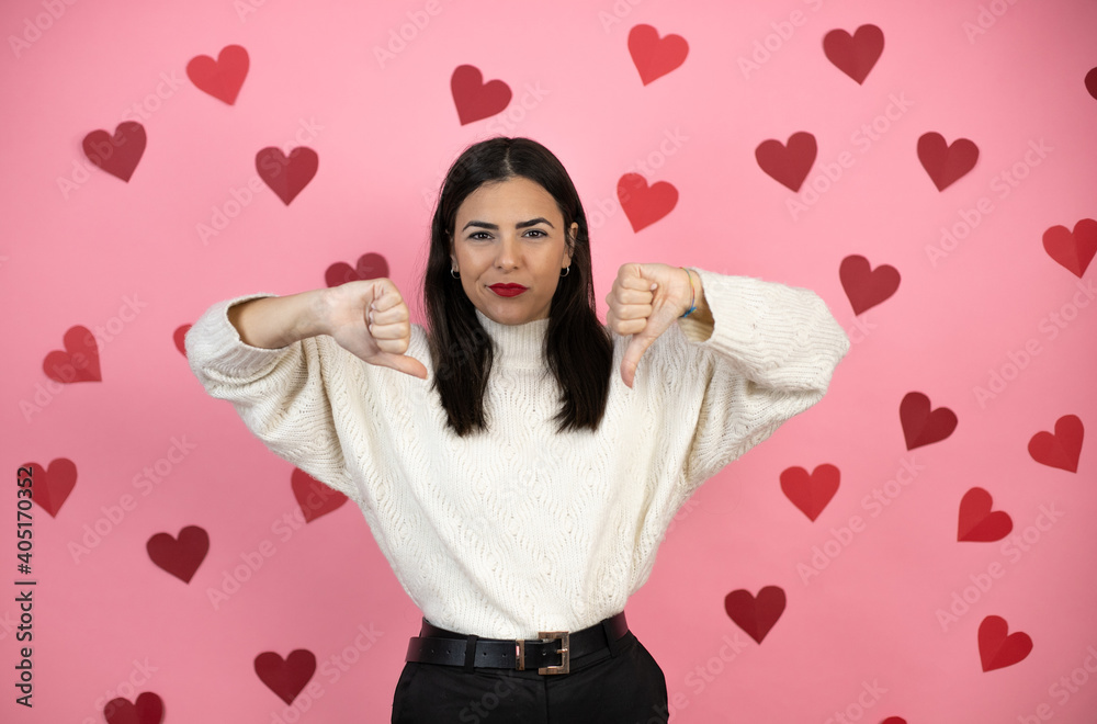 Young beautiful woman over pink background with harts with angry face, negative sign showing dislike with thumb down Young beautiful woman over pink background with harts with angry face, negative sign showing dislike with thumb down