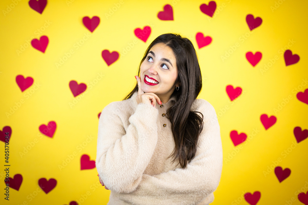 Young caucasian woman over yellow background with red hearts with hand on chin thinking about question, pensive expression. smiling and thoughtful face. doubt concept. Young caucasian woman over yellow background with red hearts with hand on chin thinking about question, pensive expression. smiling and thoughtful face. doubt concept.