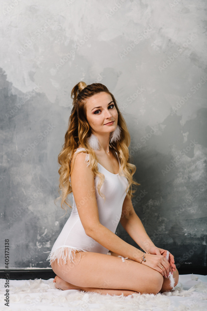 Beautiful young girl in swimsuit with feathers. Stylish woman in boho style bodysuit sits on floor on gray wall background.