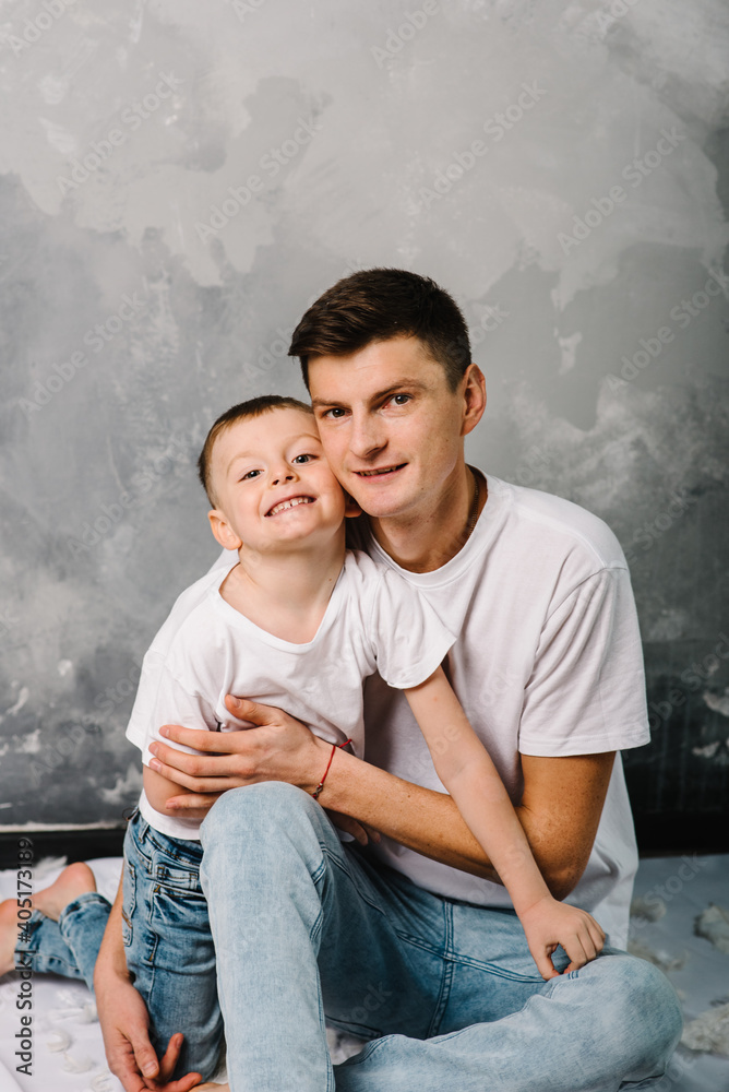 Smiling dad with son sitting on the floor. Fashion models plays with feathers. Happy family portrait in casual style clothes. Nice family wearing jeans isolated on grey wall.