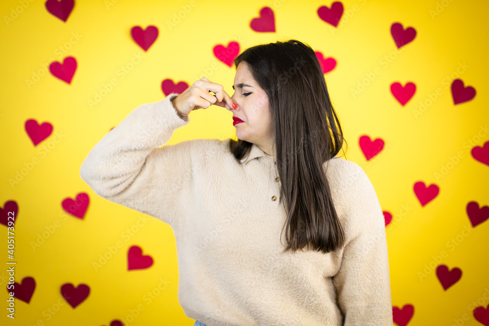 Young caucasian woman over yellow background with red hearts smelling something stinky and disgusting, intolerable smell, holding breath with fingers on nose Young caucasian woman over yellow background with red hearts smelling something stinky and disgusting, intolerable smell, holding breath with fingers on nose