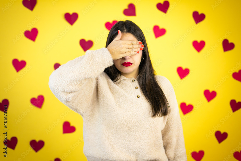 Young caucasian woman over yellow background with red hearts serious and covering her eyes with her hand Young caucasian woman over yellow background with red hearts serious and covering her eyes with her hand