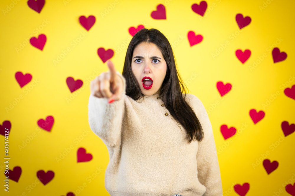 Young caucasian woman over yellow background with red hearts pointing with finger surprised ahead, open mouth amazed expression, something on the front Young caucasian woman over yellow background with red hearts pointing with finger surprised ahead, open mouth amazed expression, something on the front
