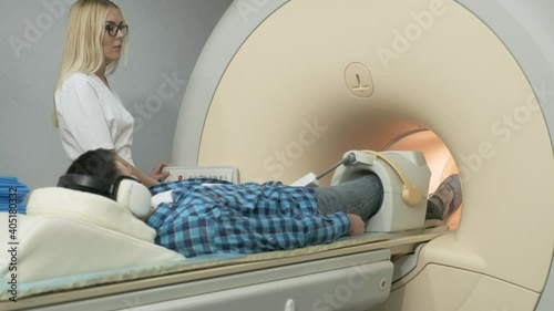 Woman doctor makes knee-joint MRI scanning. Young man patient on automatic table enters into a closed-type mri machine using noise isolation headphones. Modern equipment, coil on the patient's knee