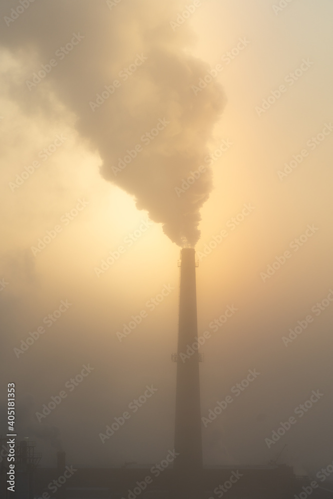 The chimney of a steaming power plant emerges from the back fog. The chimney of the power plant rises above the surrounding fog. Energy. Heating plant in operation. The chimney of a steaming power plant emerges from the back fog. The chimney of the power plant rises above the surrounding fog. Energy. Heating plant in operation.