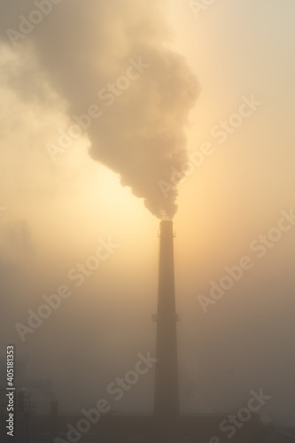 The chimney of a steaming power plant emerges from the back fog. The chimney of the power plant rises above the surrounding fog. Energy. Heating plant in operation.