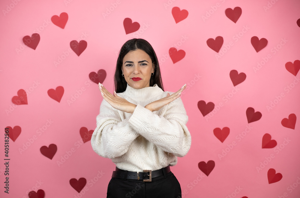Young beautiful woman over pink background with harts Rejection expression crossing arms doing negative sign, angry face Young beautiful woman over pink background with harts Rejection expression crossing arms doing negative sign, angry face