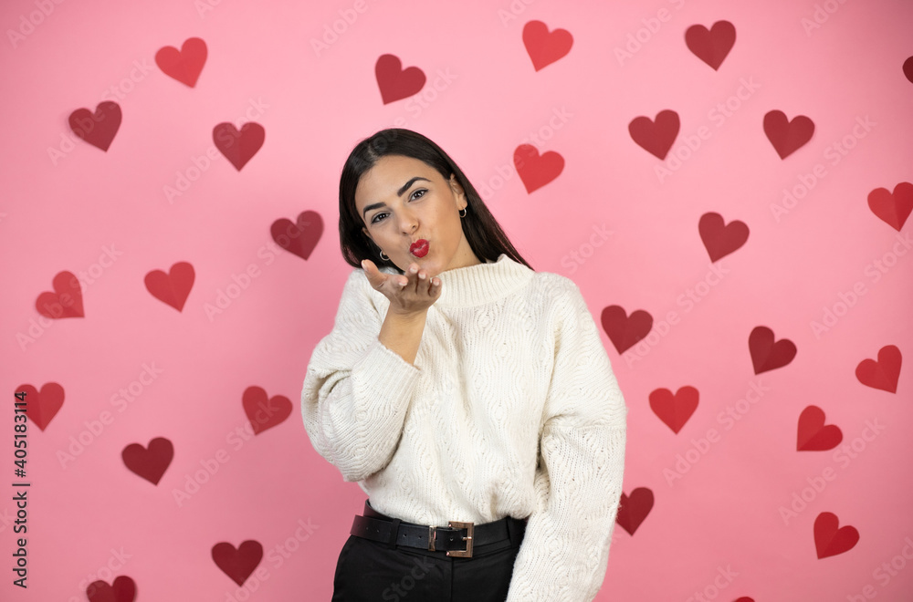 Young beautiful woman over pink background with harts looking at the camera blowing a kiss with hand on air being lovely and sexy. Love expression. Young beautiful woman over pink background with harts looking at the camera blowing a kiss with hand on air being lovely and sexy. Love expression.