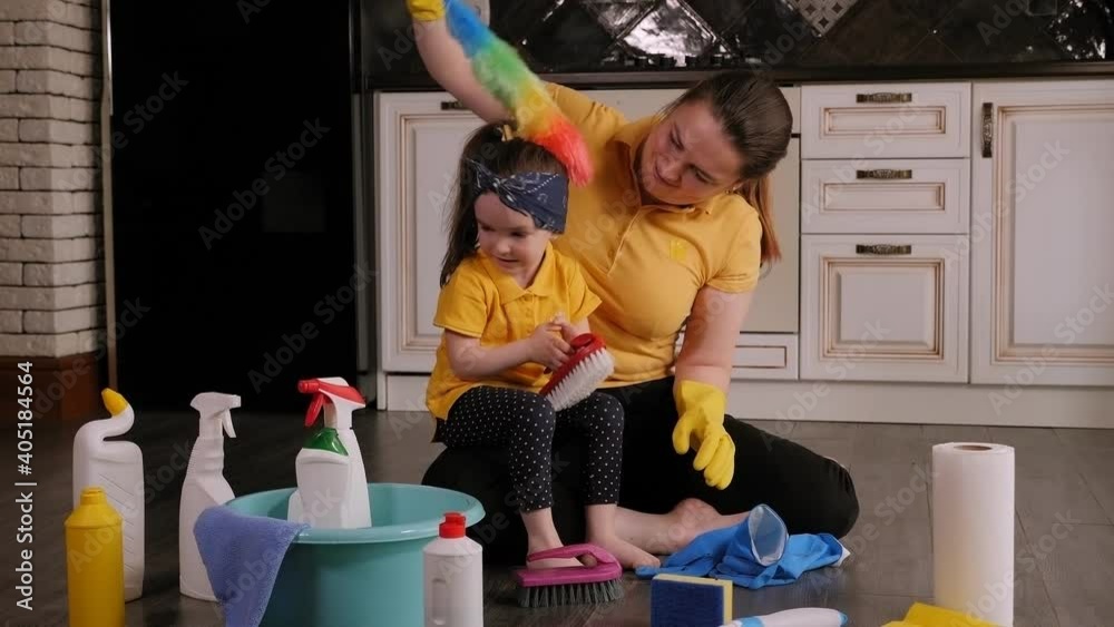 A happy mother with a small charming daughter is preparing to clean the house, they sit on the floor among detergents, holding brushes in their hands and laughing. A happy mother with a small charming daughter is preparing to clean the house, they sit on the floor among detergents, holding brushes in their hands and laughing.
