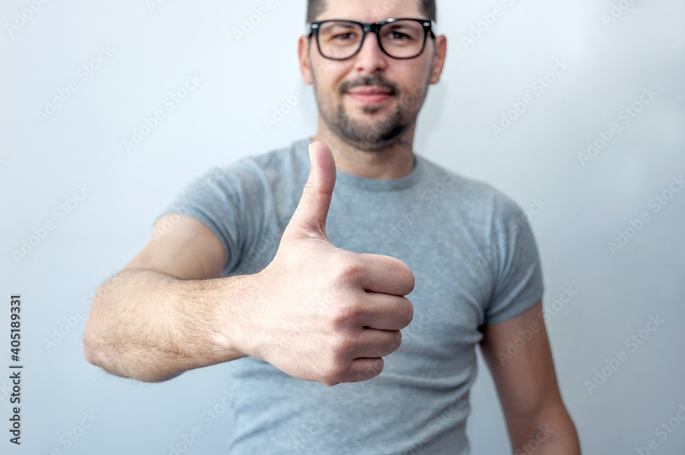 Happy young caucasian man in a gray t-shirt makes a thumb up sign showing his support, respect and recommended for someone. Language of the body. I like it. Well done. Promotion. Public relations. Happy young caucasian man in a gray t-shirt makes a thumb up sign showing his support, respect and recommended for someone. Language of the body. I like it. Well done. Promotion. Public relations.