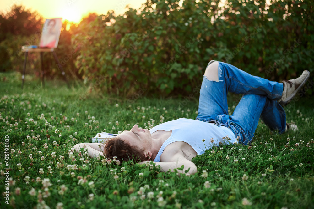 Young male artist, wearing torn jeans and white t-shirt, lying on green grass during sunset, with empty canvas beside, thinking what to draw. Painting workshop outside. Artistic inspiration concept. Young male artist, wearing torn jeans and white t-shirt, lying on green grass during sunset, with empty canvas beside, thinking what to draw. Painting workshop outside. Artistic inspiration concept.