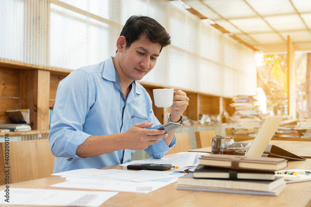 Businessman using smartphone and holding coffee cup while working on laptop.