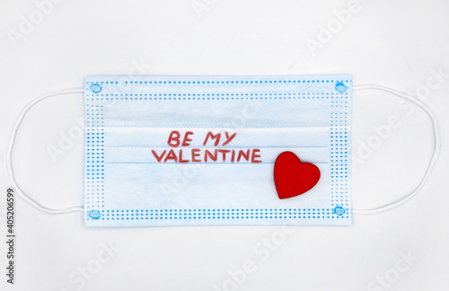 Top view Medical protective face mask with red heart shape and text BE MY VALENTINE on white background. Valentines Day concept, stay safe. Flatlay with message photo