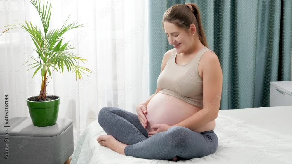 SMiling relaxed pregnant woman stroking and touching her belly against big window in bedroom. Concept of healthy lifestyle, healthcare and sports during pregnancy SMiling relaxed pregnant woman stroking and touching her belly against big window in bedroom. Concept of healthy lifestyle, healthcare and sports during pregnancy
